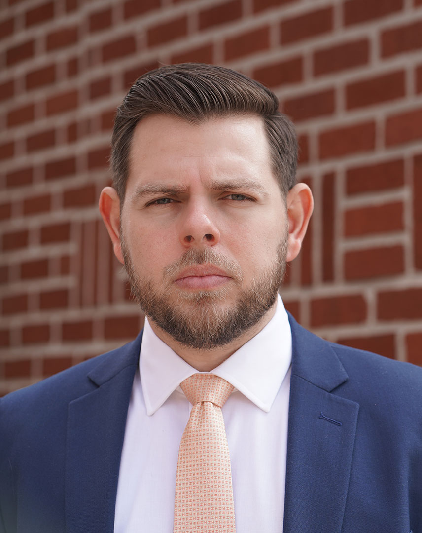 Portrait of a man in a navy suit and peach tie standing against a red brick wall.
